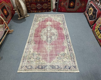 May include: A rectangular, distressed-look rug with a central medallion design. The rug features a faded, reddish-pink field with intricate floral and geometric patterns in cream, navy, and light blue. The rug is surrounded by other decorative rugs and pillows.