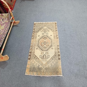 Puede incluir: Una alfombra rectangular de estilo vintage con un diseño geométrico central en tonos beige, marrón y naranja. La alfombra tiene un aspecto envejecido, con colores descoloridos y una textura desgastada, lo que sugiere que está hecha a mano.