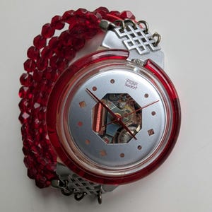 Puede incluir: Un reloj Swatch POP rojo y plateado con una esfera esqueletizada y una pulsera de cuentas rojas.