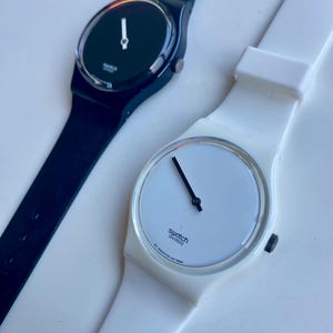 May include: Two wristwatches with white straps. The watch on the left has a black face and the watch on the right has a white face. Both watches have black hands and say "Swatch Swiss" on the face.