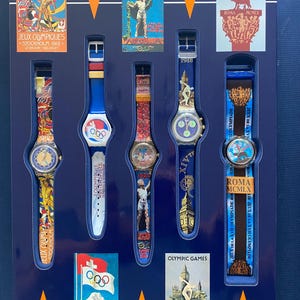 Puede incluir: Una colección de relojes de pulsera vintage con temática de los Juegos Olímpicos. Cada reloj presenta diseños y colores únicos, con correas que muestran símbolos olímpicos, nombres de ciudades y años de eventos. La exhibición incluye carteles históricos y texto relacionado.