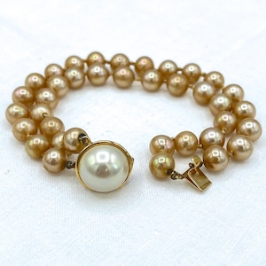 May include: A double-strand bracelet featuring lustrous, golden-toned pearls. The bracelet has a large, off-white pearl as a clasp, set in a gold-colored metal frame. The clasp is connected to a gold-colored metal clasp.