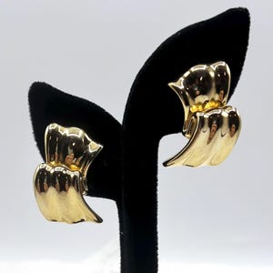 May include: A pair of gold-toned clip-on earrings. Each earring features a layered, abstract design with a curved, wing-like shape. The earrings are displayed on a black velvet earring stand against a white background.