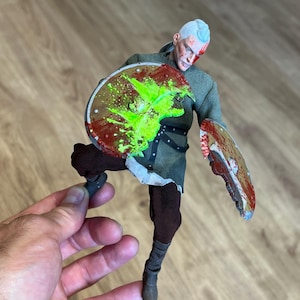 Op de afbeelding: Gedetailleerde actiefiguur van een krijger in een groene tuniek en bruine broek, met twee schilden. De schilden zijn bespat met rode en groene verf, wat wijst op een gevechtsscène. De figuur heeft wit haar en een bebloed gezicht.