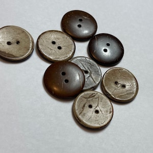 Könnte beinhalten: Acht braune und beige Kokosnuss-Knöpfe mit jeweils zwei Löchern. Die Knöpfe sind in einem Halbkreis auf einer weißen Oberfläche angeordnet.