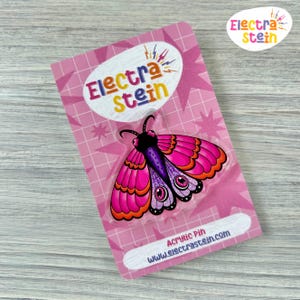 Op de afbeelding: Een roze acrylspeld met een gestileerd motontwerp met levendige roze, paarse en oranje vleugels. De speld zit op een roze kaart met de tekst "Electra Stein" en "Acrylic Pin www.electrastein.com".