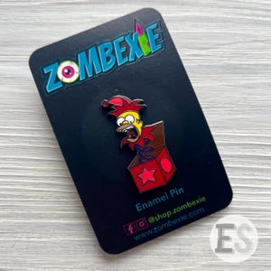 May include: A yellow cartoon character with a red jester hat pops out of a red and brown box with a white star. The enamel pin is on a black card with the text "Zombexie" and "Enamel Pin".