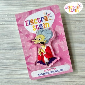 May include: An acrylic pin depicting a cartoon character in a red jacket and blue hair, with an open mouth. The pin is on a pink backing with the text "Electra Stein" and "Acrylic Pin www.electrastein.com". The background has a star pattern.