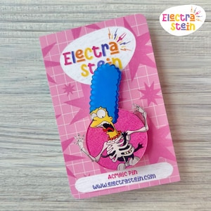 May include: A pink and white card with the text "Electra Stein" in a pink and yellow font. The card features a cartoon image of a yellow character with blue hair and a skeleton body. The character is wearing a yellow necklace and has a shocked expression. The text "Acrylic Pin" and "www.electraste.com" are printed on the card.