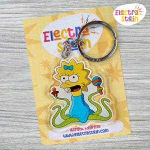 Może przedstawiać: Żółte tło z białym wzorem gwiazd. Przezroczysty brelok z akrylu z kreskówkową postacią dziewczynki o żółtych włosach, niebieskich oczach i niebieskiej sukience. Dziewczynka trzyma grzechotkę i ma smoczek w ustach. Brelok jest przymocowany do srebrnego kółka na klucze. Tekst "Electra Stein" jest nadrukowany na tle w kolorach tęczy.