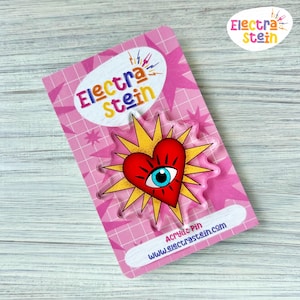 May include: A clear acrylic pin featuring a red heart with a blue eye design, surrounded by yellow rays. The pin is on a pink backing card with the text "Electra Stein" and "Acrylic Pin www.electrastein.com".