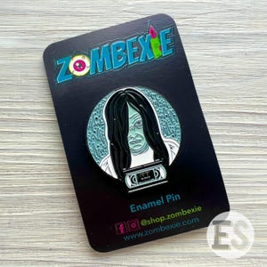 Peut inclure: Épinglette en émail noir représentant une femme aux longs cheveux noirs et une chemise blanche, tenant une cassette VHS. L'épingle est sur une carte noire avec le texte "ZOMBEXIE" en bleu et blanc, et "Enamel Pin" en blanc. La carte comprend également le texte "@shop.zombexie" et "www.zombexie.com" en blanc.