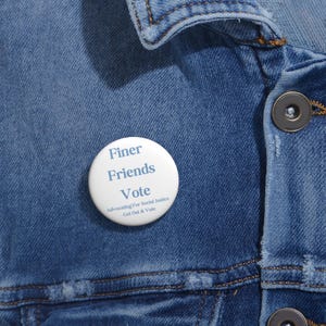 May include: A white button with the words "Finer Friends Vote" in light blue, along with the text "Advocating For Social Justice Get Out & Vote." The button is pinned to a blue denim jacket.