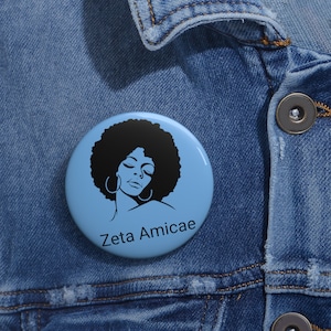 May include: A light blue button with a black illustration of a woman with an afro and hoop earrings. The text "Zeta Amicae" is printed in black below the illustration. The button is round and appears to be made of metal or plastic.