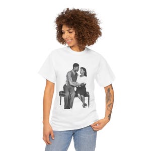 May include: White t-shirt featuring a black and white image of a couple sitting on a bench, embracing. The image is a vintage photograph. The t-shirt is being worn by a person with curly brown hair and blue jeans.