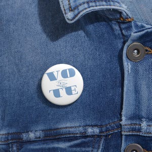 May include: A white button with the word "VOTE" in light blue block letters. The button is on a blue denim jacket. The button also has a light blue graphic of two hands shaking.