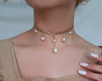 Könnte beinhalten: Goldfarbene Choker-Halskette mit hängenden Kunstperlen. Die Halskette hat ein filigranes Kettendesign mit kleinen Perlen und einer größeren tropfenförmigen Perle. Ein stilvolles Accessoire.