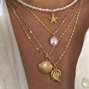 Op de afbeelding: Een gelaagde ketting set met gouden kettingen en hangers. De bovenste laag is een parelketting met een zeester bedel. Daaronder zijn gouden kettingen met een parel, een zeeschelp en een spiraalvormige hanger.