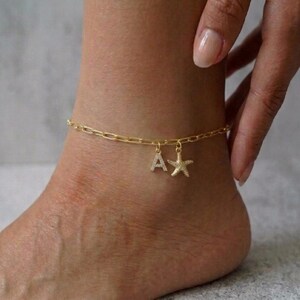 Op de afbeelding: Goudkleurige enkelband met een schakelkettingontwerp. De enkelband heeft twee bedels: de letter "A" en een zeester, beide versierd met kleine, sprankelende versieringen. De enkelband wordt om een enkel gedragen.