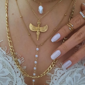 Op de afbeelding: Een verzameling gouden kettingen met verschillende ontwerpen, waaronder een schakelketting, een hanger met een parel en een hanger met een gevleugelde figuur. De kettingen zijn gelaagd en worden samen gedragen.