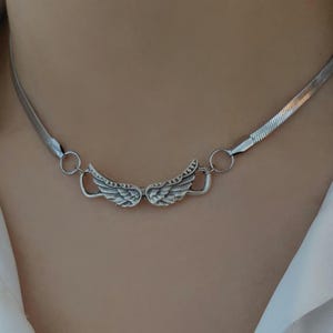 Op de afbeelding: Een zilverkleurige ketting met een paar gedetailleerde vleugelbedels. De ketting heeft een plat, slangenkettingontwerp en wordt vastgezet met een eenvoudige sluiting. De vleugels zijn ingewikkeld ontworpen.