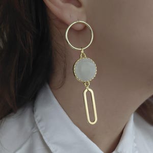 Op de afbeelding: Goudkleurige oorbellen met een cirkelvormige bovenkant, een ronde, lichtgekleurde steen en een hangende ovale vorm. De oorbellen worden gedragen door een persoon die een wit overhemd draagt.