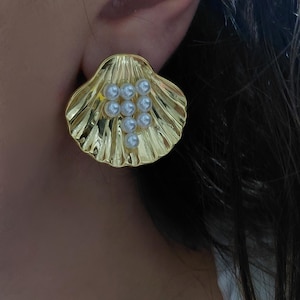 Op de afbeelding: Goudkleurige oorknopjes in de vorm van een zeeschelp met een cluster van kleine, witte parels. De oorbellen hebben een getextureerd oppervlak en worden op het oor gedragen. Een modeaccessoire.