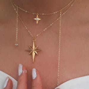 Peut inclure: Ensemble de colliers dorés superposés. Il comprend un pendentif en forme d'étoile filante, un petit pendentif en forme d'étoile avec une pierre transparente et une chaîne avec un charme en forme de goutte. Les bijoux sont portés sur la poitrine.