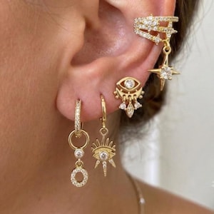 May include: A set of four gold-toned earrings featuring clear cubic zirconia stones. The collection includes a star-shaped ear cuff, an eye-shaped stud with dangling charms, a sunburst charm hoop, and a hoop earring with a circular drop pendant.