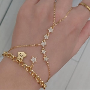 Op de afbeelding: Een goudkleurige handketting bestaande uit een fijne schakelarmband verbonden met een ring via een ketting versierd met vijf stervormige bedels ingelegd met heldere steentjes. Een hartvormige bedel met de letter S hangt aan de armband.