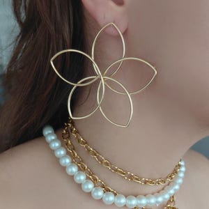 Op de afbeelding: Goudkleurige bloemvormige oorbellen en een gelaagde ketting met parels en een gouden ketting. De oorbellen zijn groot en hebben een delicaat, open ontwerp. De ketting heeft meerdere strengen.
