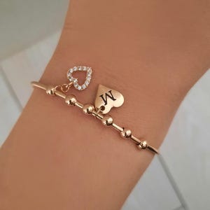 Op de afbeelding: Een roségoudkleurige armband met een reeks kleine, ronde kralen langs de band. De armband bevat twee bungelende bedels: een hartvormige bedel ingelegd met heldere steentjes en een tweede hartvormige bedel gegraveerd met de letter M.