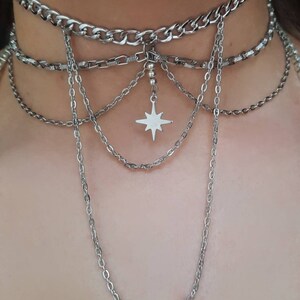K&ouml;nnte beinhalten: Eine mehrreihige silberne Kettenkette mit einem Sternanh&auml;nger. Die Kette hat oben eine dicke Kette und mehrere d&uuml;nnere Ketten, die herunterh&auml;ngen.
