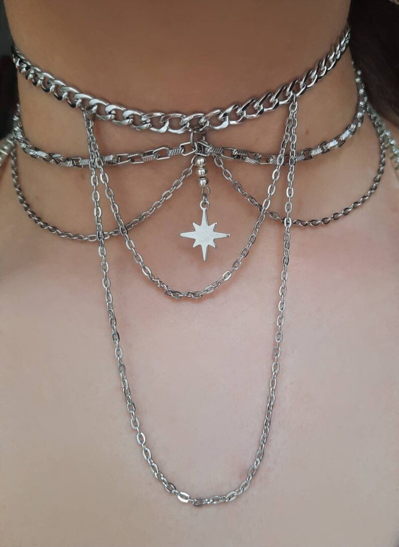 K&ouml;nnte beinhalten: Eine mehrreihige silberne Kettenkette mit einem Sternanh&auml;nger. Die Kette hat oben eine dicke Kette und mehrere d&uuml;nnere Ketten, die herunterh&auml;ngen.