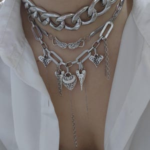 Op de afbeelding: Een gelaagde zilveren ketting set. De bovenste laag is een dikke ketting, gevolgd door een ketting met vleugelbedels en een ketting met hartbedels. De harten hebben verschillende texturen en vormen, met bungelende kettingen.