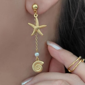 Op de afbeelding: Een goudkleurige hangende oorbel met een getextureerde zeesterbedel aan een kettinkje, geaccentueerd met een kleine witte parel en een spiraalvormige schelpbedel aan de onderkant, bevestigd met een goudkleurige bolvormige oorsteker.