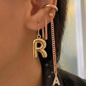 Op de afbeelding: Een set goudkleurige oorbellen met een gladde, opgeblazen letter R-bedel, een rechthoekige staaf-oorbel, een eenvoudige oormanchet en een lange ketting-oorbel met een kleine, met kristallen ingelegde letter A-hanger aan de onderkant.