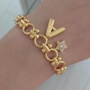 Op de afbeelding: Een goudkleurige schakelarmband met een grote, gladde letter A-bedel en een kleinere, vijfpuntige sterbedel bezet met heldere, fonkelende steentjes. De armband is opgebouwd uit een reeks onderling verbonden metalen ringen.