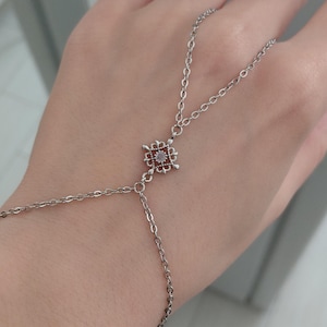 Op de afbeelding: Een zilveren handketting met een centraal vierkant ontwerp, met een kleine, ronde, lichtgekleurde steen. De ketting loopt door naar de vinger en pols, waardoor een delicaat en elegant accessoire ontstaat.