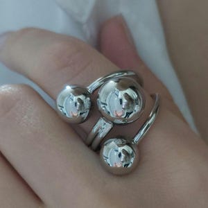 Op de afbeelding: Een zilverkleurige ring met drie bolvormige bollen. De bollen zijn verbonden door gebogen banden die de vinger omsluiten. De ring heeft een modern, minimalistisch ontwerp en weerspiegelt de omgeving.