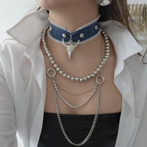 Könnte beinhalten: Halskette mit Denim-Choker, silbernen Nieten und einem Bullenschädel-Anhänger. Die Halskette hat mehrere Stränge aus silbernen Perlen und Ketten, die eine kühne, kantige Ästhetik erzeugen.