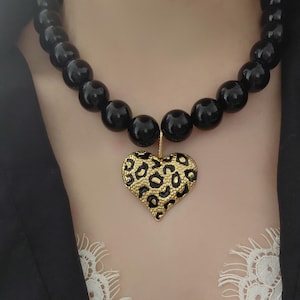 Op de afbeelding: Een zwarte kralenketting met een gouden hartvormige hanger. De hartvormige hanger heeft een luipaardprint. De ketting is een opvallend sieraad, perfect om een vleugje glamour aan elke outfit toe te voegen.