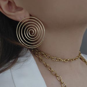 Op de afbeelding: Goudkleurige spiraalvormige oorbel. De oorbel heeft een grote, cirkelvormige vorm met concentrische cirkels die een spiraal vormen. Een gouden ketting is ook zichtbaar. De sieraden worden gedragen met een witte blazer.