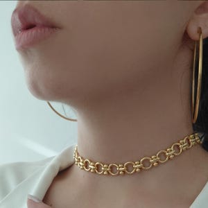 Op de afbeelding: Een goudkleurige choker-halsketting met een herhalend patroon van cirkelvormige schakels verbonden door kleine cilindrische staafjes, gecombineerd met grote, dunne, cirkelvormige goudkleurige oorringen. De sieraden hebben een gepolijste, glanzende afwerking.