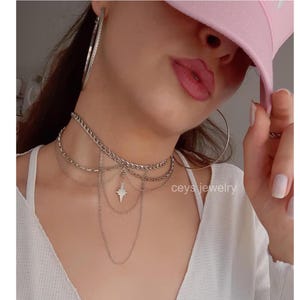 K&ouml;nnte beinhalten: Eine mehrreihige silberne Choker-Kette mit einem Sternanh&auml;nger. Die Kette wird von einer Person mit einem rosafarbenen Hut getragen.