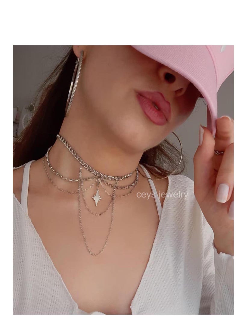 K&ouml;nnte beinhalten: Eine mehrreihige silberne Choker-Kette mit einem Sternanh&auml;nger. Die Kette wird von einer Person mit einem rosafarbenen Hut getragen.