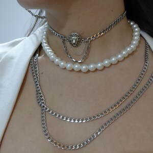 Op de afbeelding: Een gelaagde ketting set met een zilveren leeuwenhanger, een streng imitatieparels en meerdere zilveren kettingkettingen. De sieraden worden gedragen tegen een witte top.