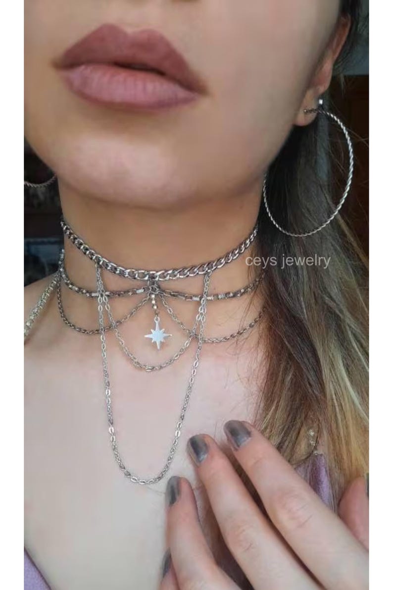 K&ouml;nnte beinhalten: Eine mehrreihige silberne Kettenkette mit einem Sternanh&auml;nger. Die Kette wird von einer Person mit dunkelgrauem Nagellack getragen. Der Text "ceys jewelry" ist im Bild sichtbar.