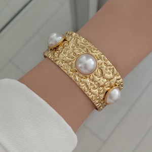 Op de afbeelding: Een brede goudkleurige manchetarmband met een getextureerd, gehamerd metalen oppervlak en drie grote, ronde, witte kunstparels die met tussenpozen in het midden van de band zijn geplaatst. De armband heeft een gepolijste afwerking.