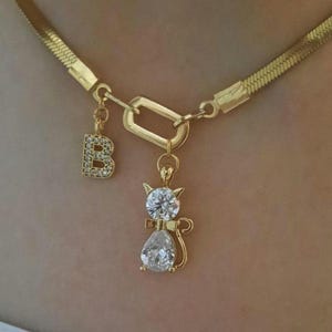 Op de afbeelding: Een goudkleurige visgraatketting met een centrale rechthoekige schakel. Aan de schakel hangen twee bedels: een letter B bezet met kleine heldere steentjes en een katvormige hanger met een ronde en een peervormige heldere steen.